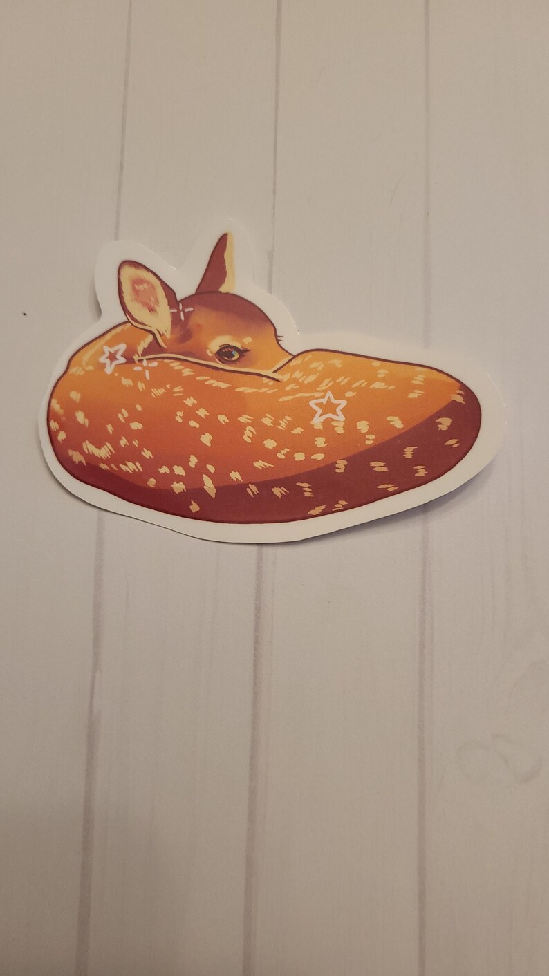 May include: A sticker featuring a curled-up fawn with a light brown coat and white spots. The fawn has a small head with visible ears and a dark eye. Two white star shapes are on the fawn's body. The sticker has a white border.