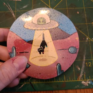 May include: A round sticker with a black cat being abducted by a UFO. The sticker has a pink, blue, and yellow background with a glittery finish. The cat has a surprised expression on its face. The UFO is white with a green alien inside.