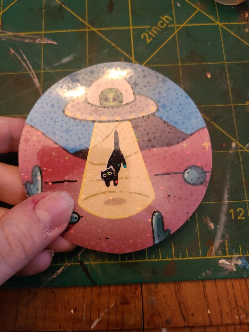 May include: A round sticker with a black cat being abducted by a UFO. The sticker has a pink, blue, and yellow background with a glittery finish. The cat has a surprised expression on its face. The UFO is white with a green alien inside.