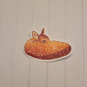 May include: A sticker featuring a sleeping fawn. The fawn is curled up, with its head tucked in. The sticker has a white border and is orange with white spots and star accents.