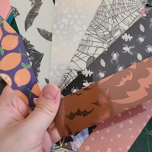May include: A collection of eight different patterned bookmarks. The bookmarks are made of paper and feature various designs, including bats, spiders, lemons, and floral patterns. The bookmarks are arranged in a fan shape and are held in a hand.