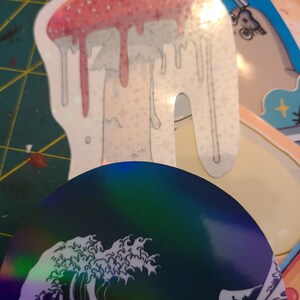 K&ouml;nnte beinhalten: Ein holografischer Aufkleber eines gro&szlig;en, tropfenden Pilzes mit einem rosa-wei&szlig;en Design. Der Aufkleber befindet sich auf einer gr&uuml;n-gelb gemusterten Oberfl&auml;che.