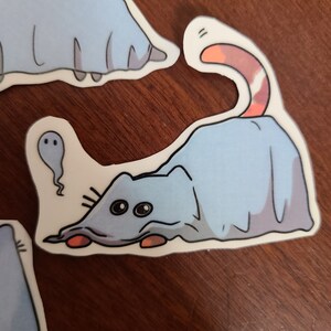 May include: A blue cat wearing a ghost costume with a white sheet and a pink tail. The cat is looking to the left with big eyes. The cat is on a white background.