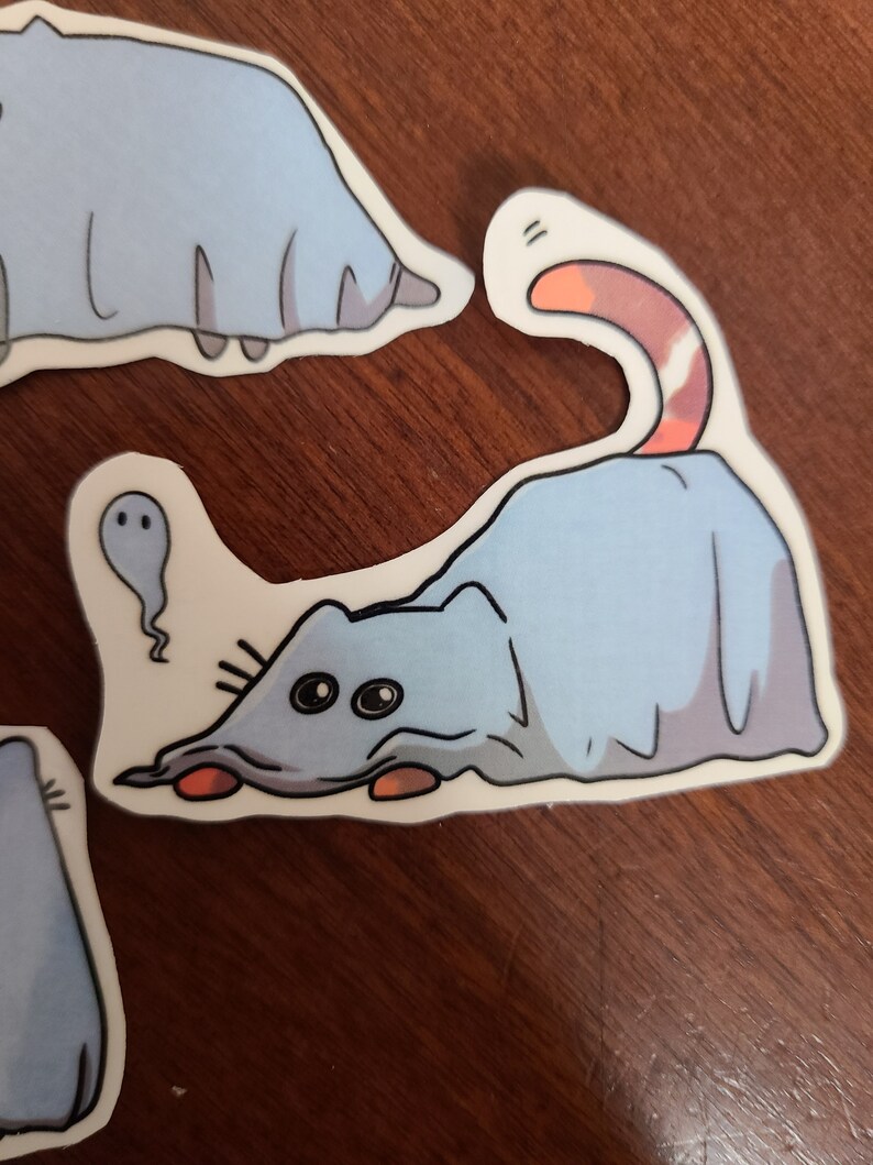 May include: A blue cat wearing a ghost costume with a white sheet and a pink tail. The cat is looking to the left with big eyes. The cat is on a white background.