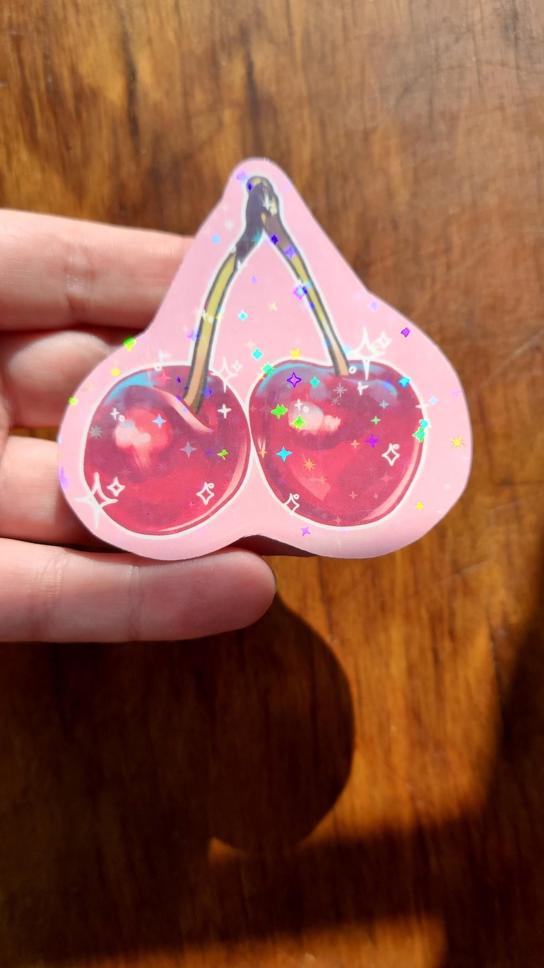 May include: A pink holographic sticker with two red cherries with green stems and leaves. The cherries are surrounded by iridescent glitter.