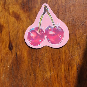 May include: A pink sticker with a pair of red cherries with shiny glitter accents.