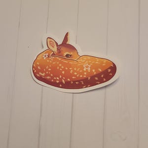 May include: A sticker featuring a stylized illustration of a sleeping fawn. The fawn is depicted in shades of brown and orange with white spots and star accents. The sticker is on a white background.