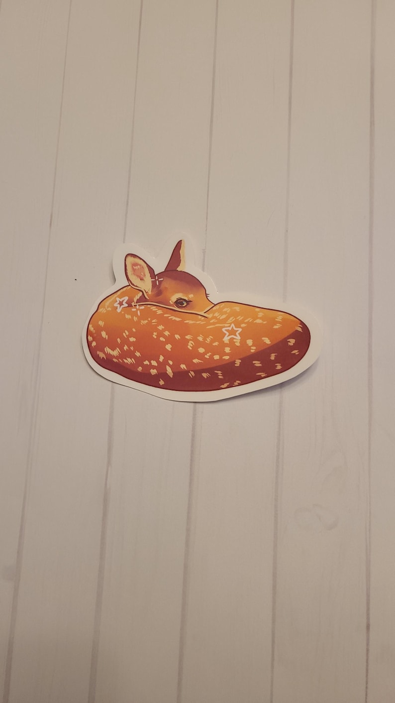 May include: A sticker featuring a stylized illustration of a sleeping fawn. The fawn is depicted in shades of brown and orange with white spots and star accents. The sticker is on a white background.