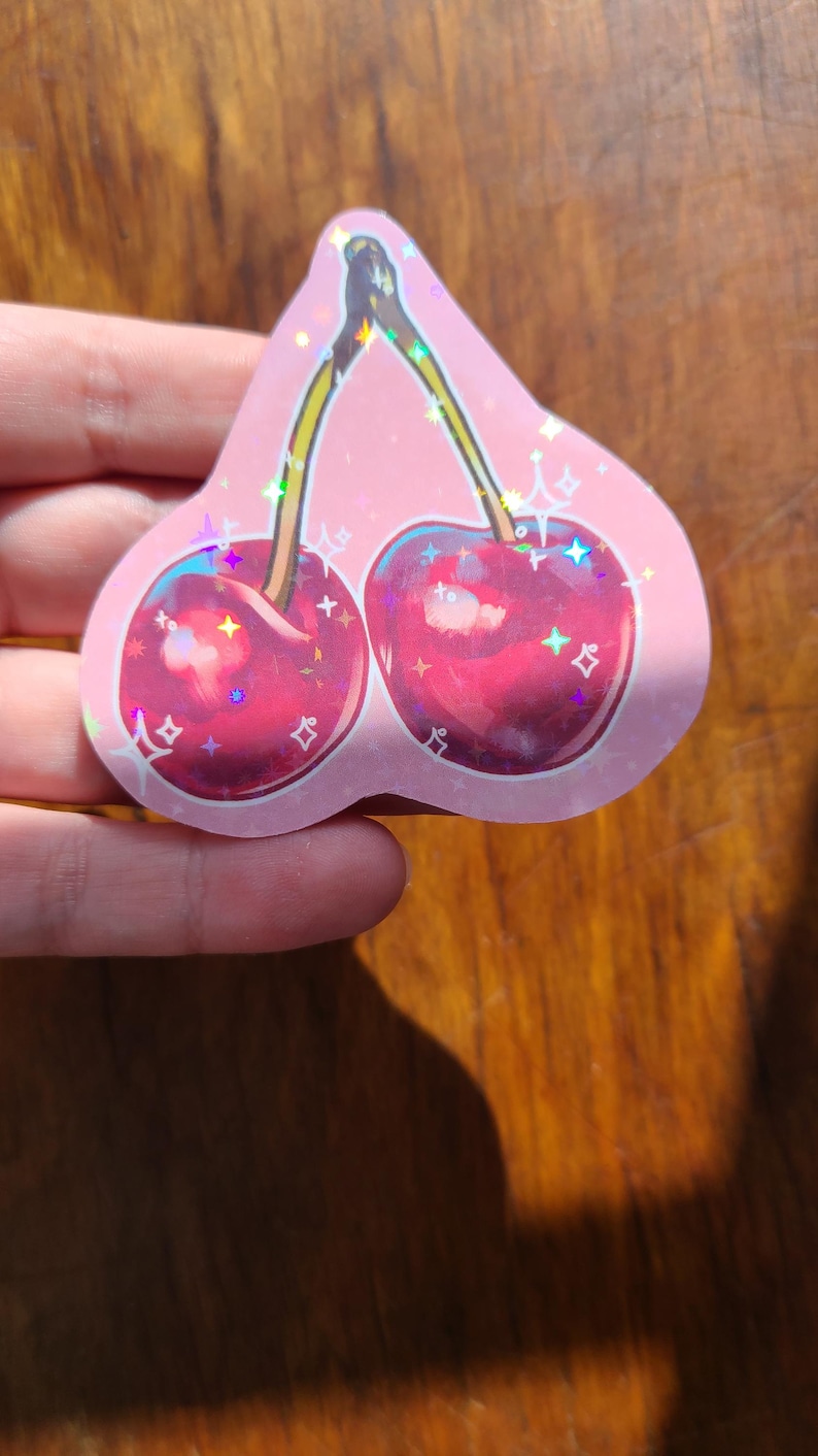 May include: A pink holographic sticker with a pair of red cherries with green stems and sparkly accents.
