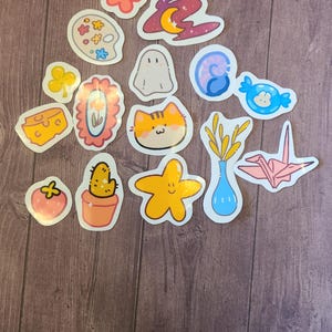 Op de afbeelding: Een verzameling van 14 kleurrijke stickers met verschillende schattige illustraties, waaronder een bloem, een geest, een kat, een cactus, een ster, een vaas met tarwe, een papieren kraanvogel, een stuk kaas, een palet, een werveling, een snoepje, een klavertjevier, een fotolijst en een aardbei.