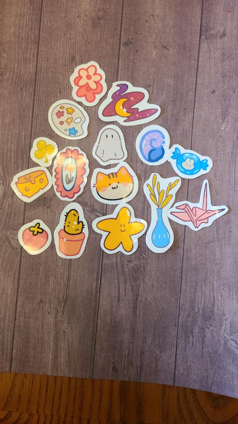 Op de afbeelding: Een verzameling van 14 kleurrijke stickers met verschillende schattige illustraties, waaronder een bloem, een geest, een kat, een cactus, een ster, een vaas met tarwe, een papieren kraanvogel, een stuk kaas, een palet, een werveling, een snoepje, een klavertjevier, een fotolijst en een aardbei.