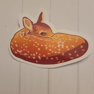 May include: A sticker featuring a curled-up fawn with a light brown coat and white spots. The fawn's head is visible, with its ears and eyes detailed. The sticker has a white border and is set against a light wood-grain background.