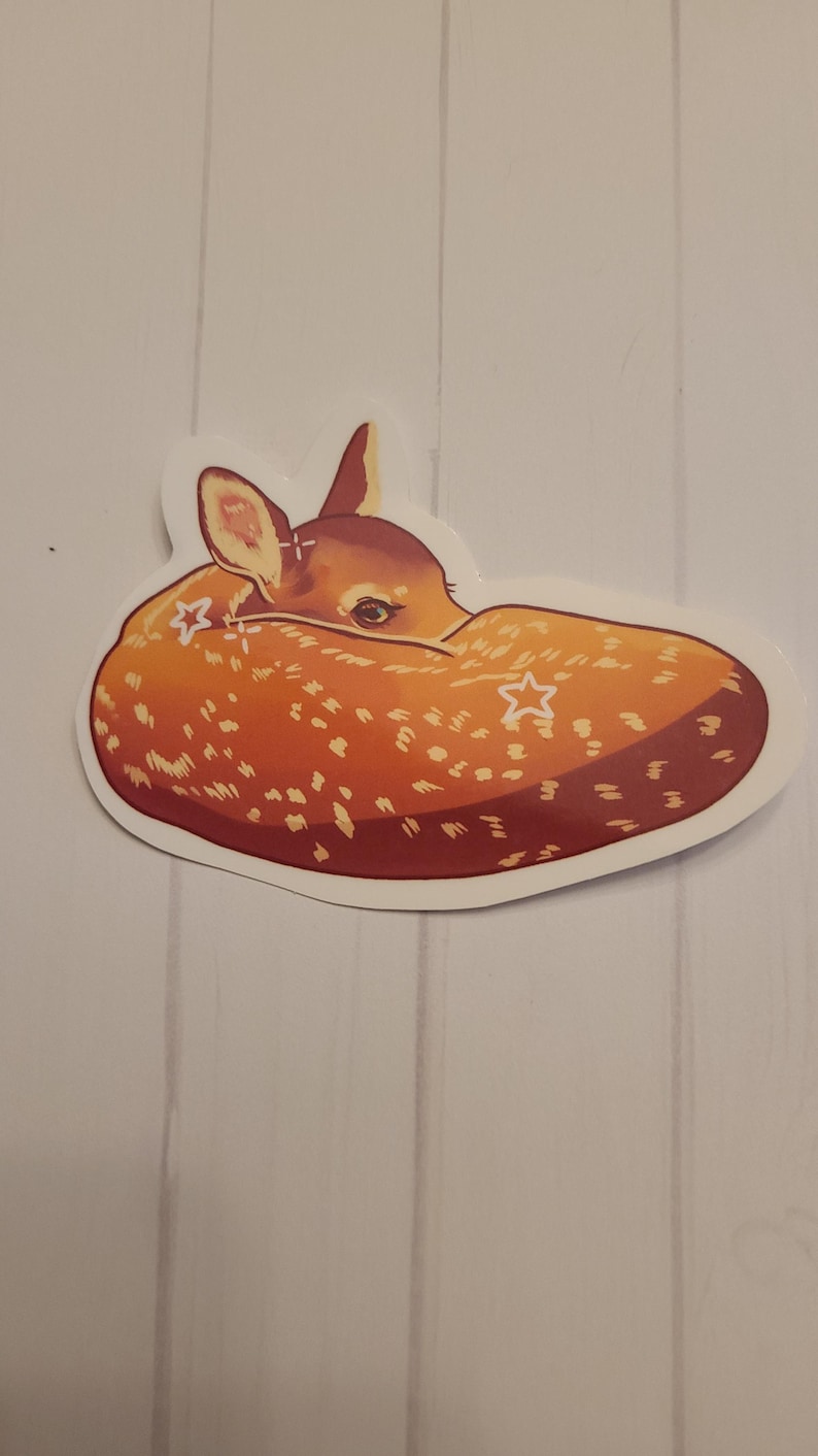 May include: A sticker featuring a curled-up fawn with a light brown coat and white spots. The fawn's head is visible, with its ears and eyes detailed. The sticker has a white border and is set against a light wood-grain background.