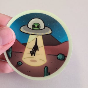 May include: A round sticker with a black cat being beamed up by a UFO. The UFO is gray with a green alien inside. The background is a dark blue night sky with stars and a desert landscape with cacti.