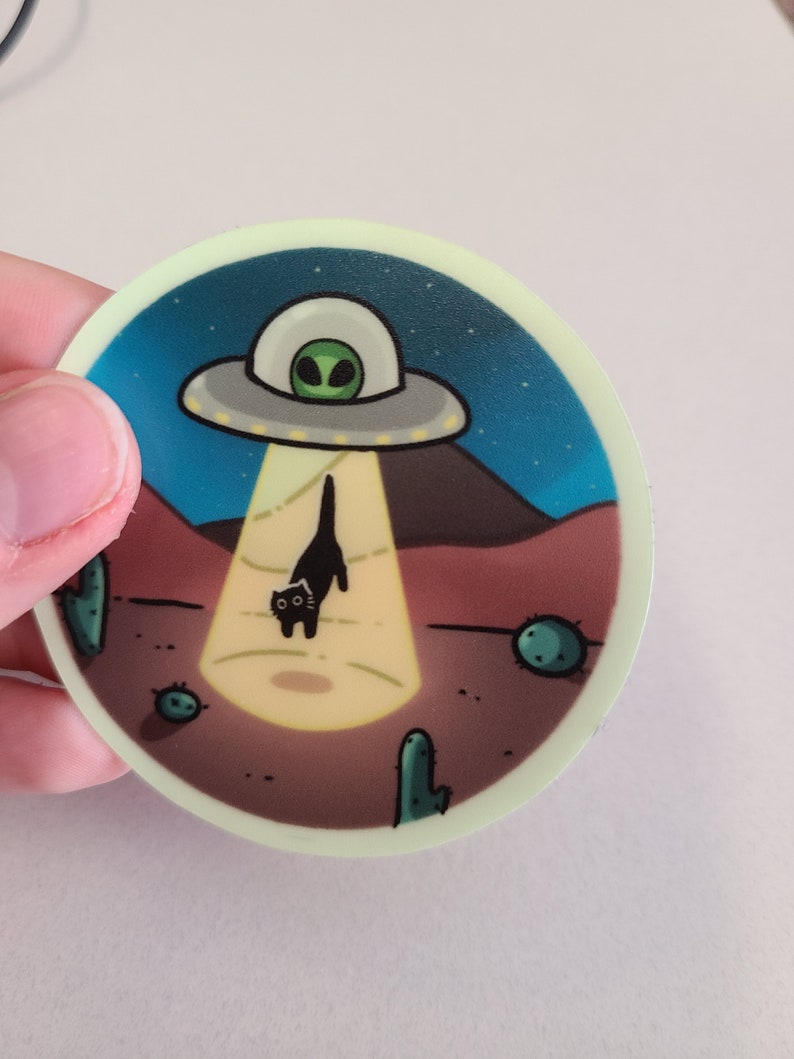 May include: A round sticker with a black cat being beamed up by a UFO. The UFO is gray with a green alien inside. The background is a dark blue night sky with stars and a desert landscape with cacti.