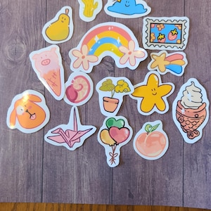 Op de afbeelding: Een verzameling kleurrijke stickers met verschillende schattige ontwerpen, waaronder een regenboog, een cactus, een wolk, een perzik, een ster, een visvormig ijsje, een plant in een pot, een origami-kraanvogel, een varken, een peer, een slak en een ballon.