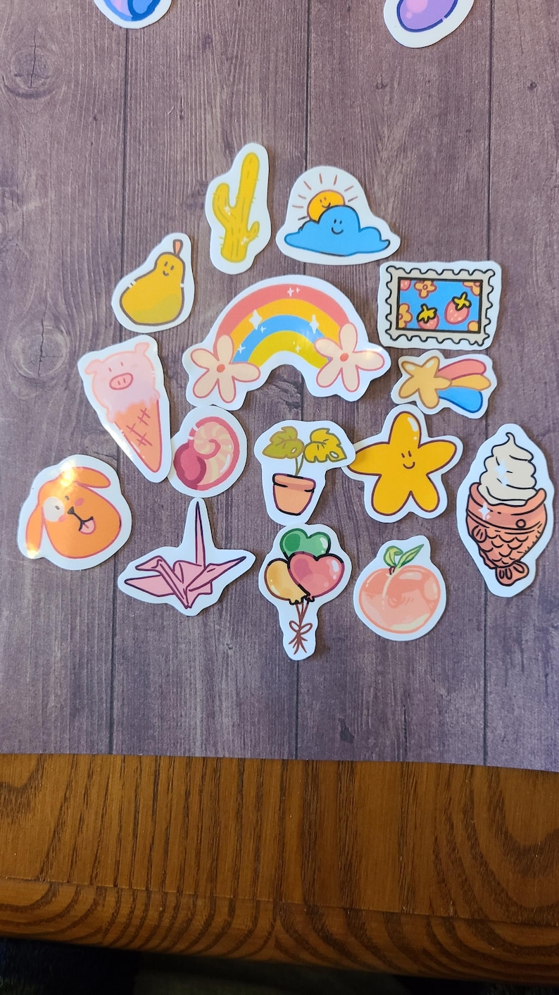 Op de afbeelding: Een verzameling kleurrijke stickers met verschillende schattige ontwerpen, waaronder een regenboog, een cactus, een wolk, een perzik, een ster, een visvormig ijsje, een plant in een pot, een origami-kraanvogel, een varken, een peer, een slak en een ballon.
