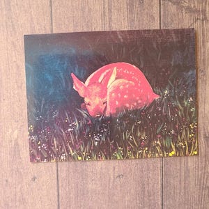 May include: A pink and white spotted fawn lying in a field of green grass and flowers. The fawn is curled up and its head is resting on its front legs.