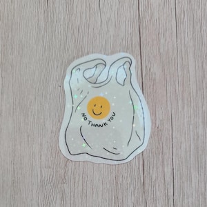 May include: A sticker featuring a white plastic bag design with a smiling yellow face in the center. The words "NO THANK YOU" are written around the face in a circular pattern. The sticker has a glittery, iridescent finish.