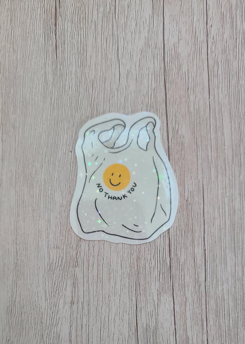 May include: A sticker featuring a white plastic bag design with a smiling yellow face in the center. The words "NO THANK YOU" are written around the face in a circular pattern. The sticker has a glittery, iridescent finish.