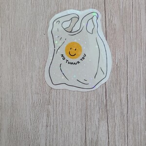 May include: A white sticker in the shape of a plastic shopping bag with the text "No thank you" and a yellow smiley face. The sticker has a holographic, glittery effect.
