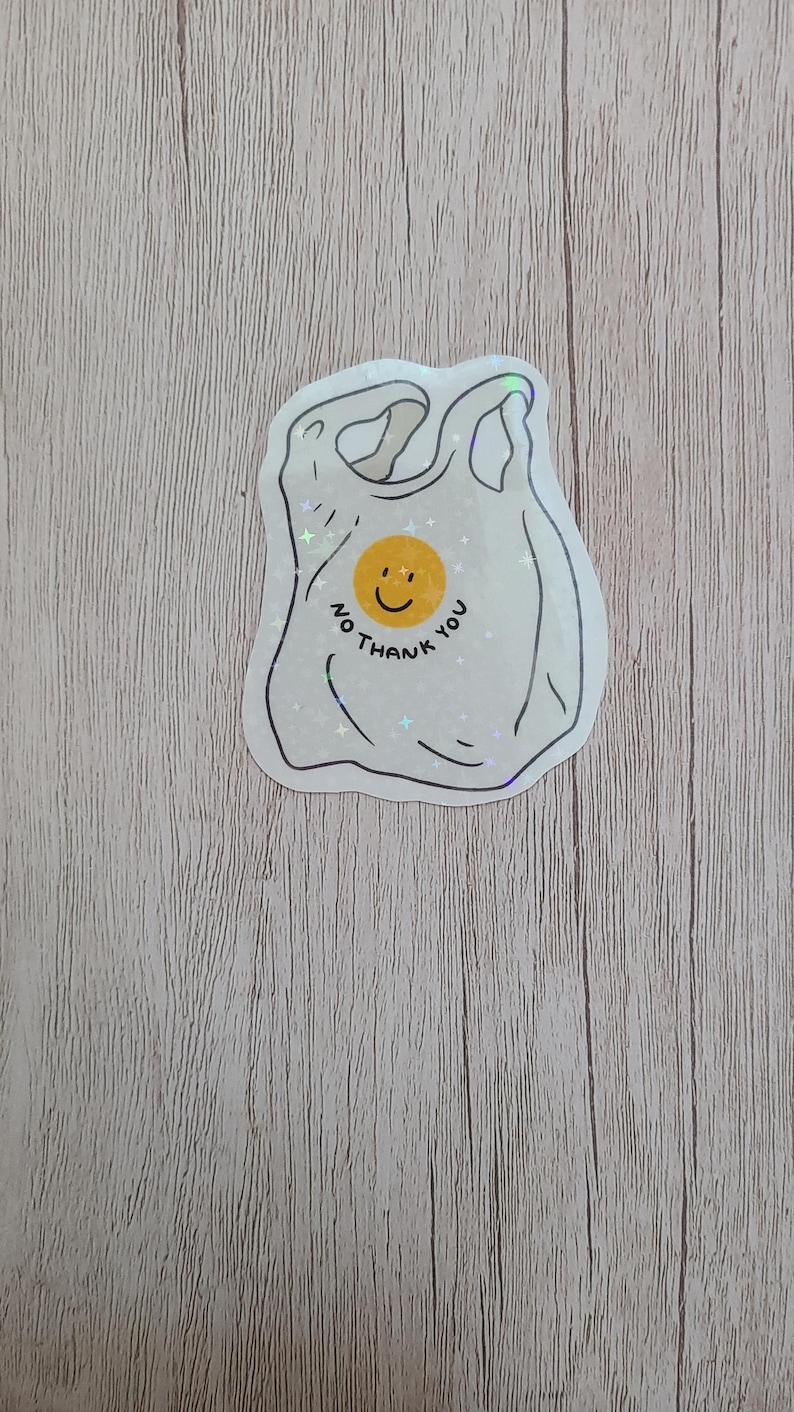 May include: A white sticker in the shape of a plastic shopping bag with the text "No thank you" and a yellow smiley face. The sticker has a holographic, glittery effect.