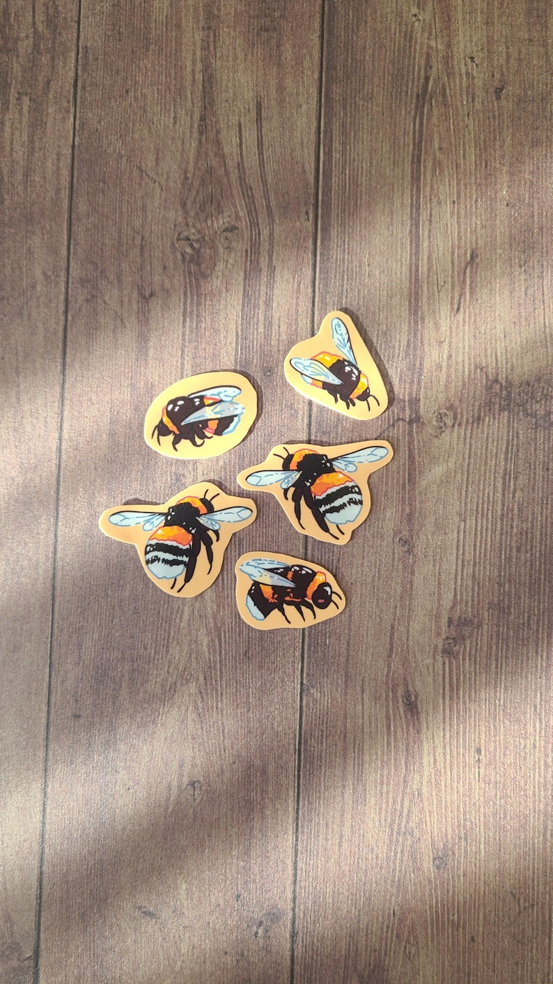 Op de afbeelding: Vijf gele en zwarte hommelstickers op een houten achtergrond. De hommels zijn in verschillende poses met hun vleugels uitgespreid.