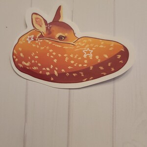 May include: A sticker featuring a curled-up fawn with a light brown coat and white spots. The fawn has a light brown head with visible ears and a dark eye. Two white star shapes are on the fawn's body. The sticker has a white border.