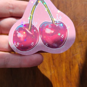 May include: A pink holographic sticker with a pair of red cherries with green stems and sparkly accents.