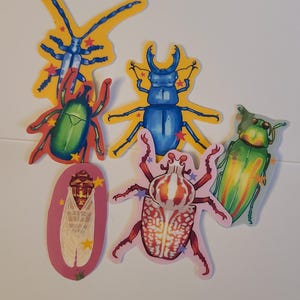 May include: A selection of insect stickers in various colours. The stickers depict beetles and cicadas in shades of blue, green, red, and pink, with yellow star accents. The stickers are arranged on a white background.