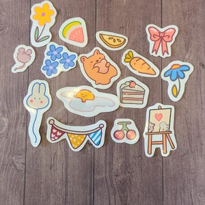 Op de afbeelding: Een set van 14 kleurrijke stickers met verschillende schattige illustraties, waaronder een konijn, een beer, een bloem, een watermeloen, een wortel, een taart, een kers, een muis, een ei, een strik, een madeliefje, een vlag en een ezel.