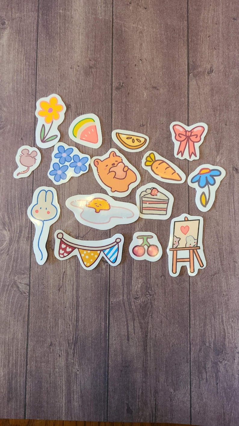 Op de afbeelding: Een set van 14 kleurrijke stickers met verschillende schattige illustraties, waaronder een konijn, een beer, een bloem, een watermeloen, een wortel, een taart, een kers, een muis, een ei, een strik, een madeliefje, een vlag en een ezel.