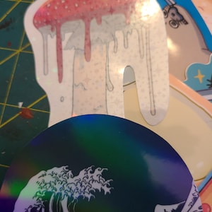 K&ouml;nnte beinhalten: Ein holografischer Aufkleber eines gro&szlig;en, tropfenden Pilzes mit einem rosa-wei&szlig;en Design. Der Aufkleber befindet sich auf einer gr&uuml;n-gelben Oberfl&auml;che.