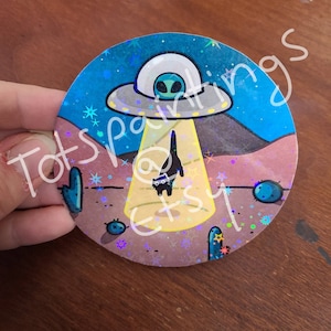 May include: A holographic sticker featuring a cartoon black cat standing in front of a UFO with a green alien inside. The UFO is emitting a beam of light. The background is a desert scene with cacti and stars.
