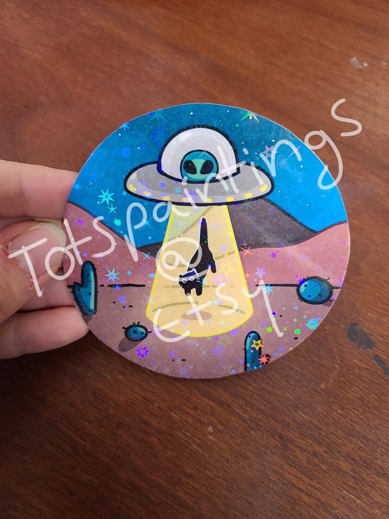 May include: A holographic sticker featuring a cartoon black cat standing in front of a UFO with a green alien inside. The UFO is emitting a beam of light. The background is a desert scene with cacti and stars.