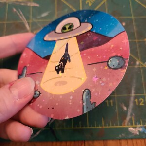 May include: A round sticker with a black cat being abducted by a UFO. The sticker has a pink, blue, and yellow background with glitter and stars. The cat has a surprised expression on its face.