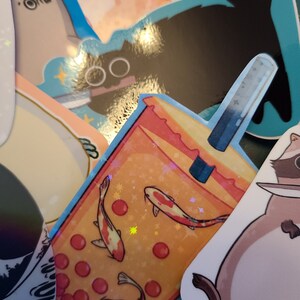 K&ouml;nnte beinhalten: Ein Aufkleber mit einem Bubble Tea mit roten und orangen Boba-Perlen, zwei Goldfischen, die im Tee schwimmen, und einem blau-wei&szlig;en Strohhalm. Der Aufkleber hat eine glitzernde Oberfl&auml;che.
