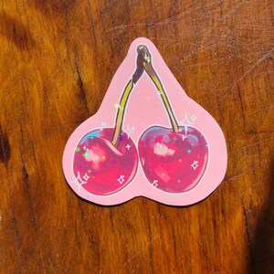 May include: A pink sticker with two red cherries with sparkly glitter accents. The cherries are drawn in a cartoon style with stems and leaves.