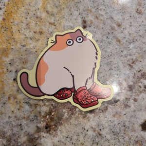 Pu&ograve; includere: Un adesivo con un gatto dei cartoni animati che indossa zoccoli rossi e gialli. Il gatto &egrave; bianco con macchie arancioni e nere. Il gatto ha grandi occhi rotondi.