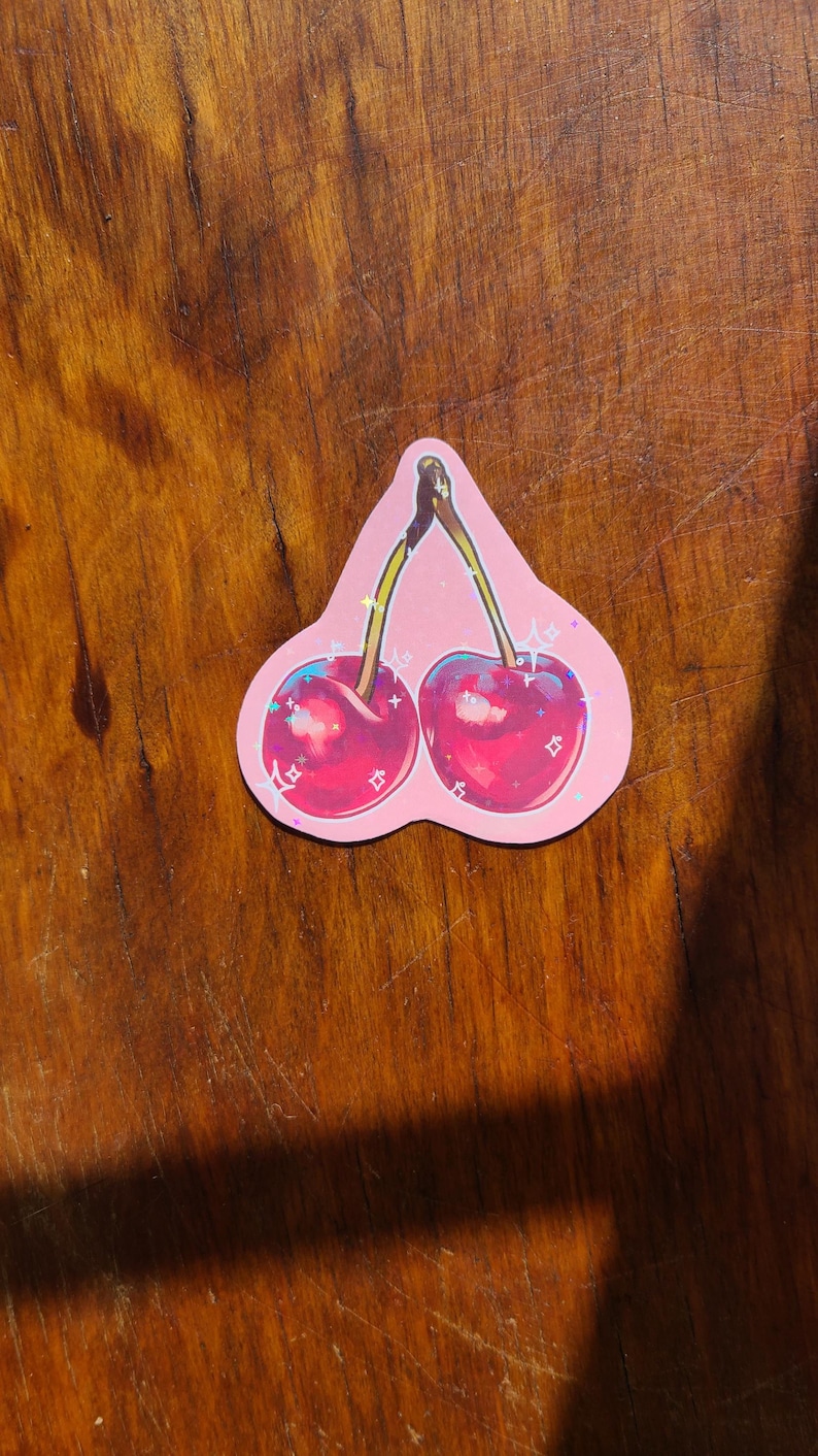 May include: A pink sticker with a pair of red cherries with green stems and leaves. The cherries have a glittery finish.