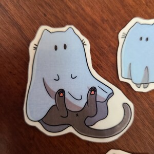 May include: A blue and gray cat wearing a ghost costume. The cat is sitting on its hind legs with its paws in the air. The ghost costume has a white outline and a black face with two eyes.
