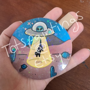 May include: A round, holographic sticker with a black cat standing in a beam of light from a UFO. The UFO is flying over a desert landscape with cacti and stars in the background.