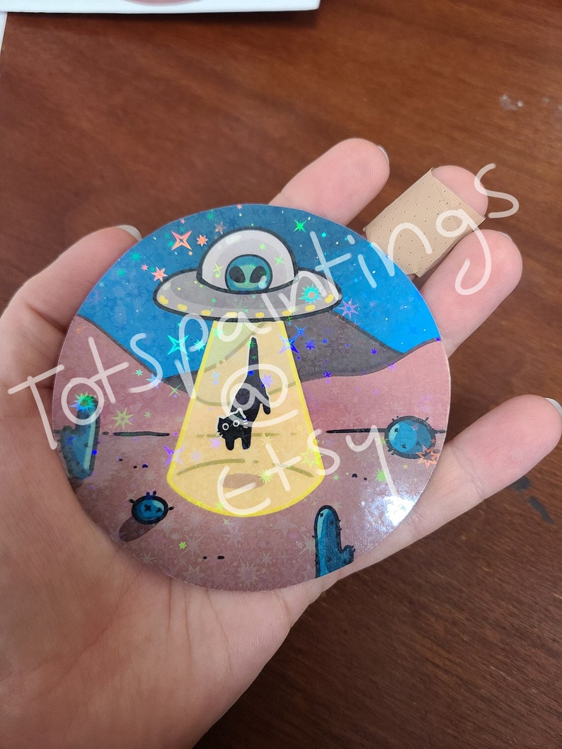 May include: A round, holographic sticker with a black cat standing in a beam of light from a UFO. The UFO is flying over a desert landscape with cacti and stars in the background.