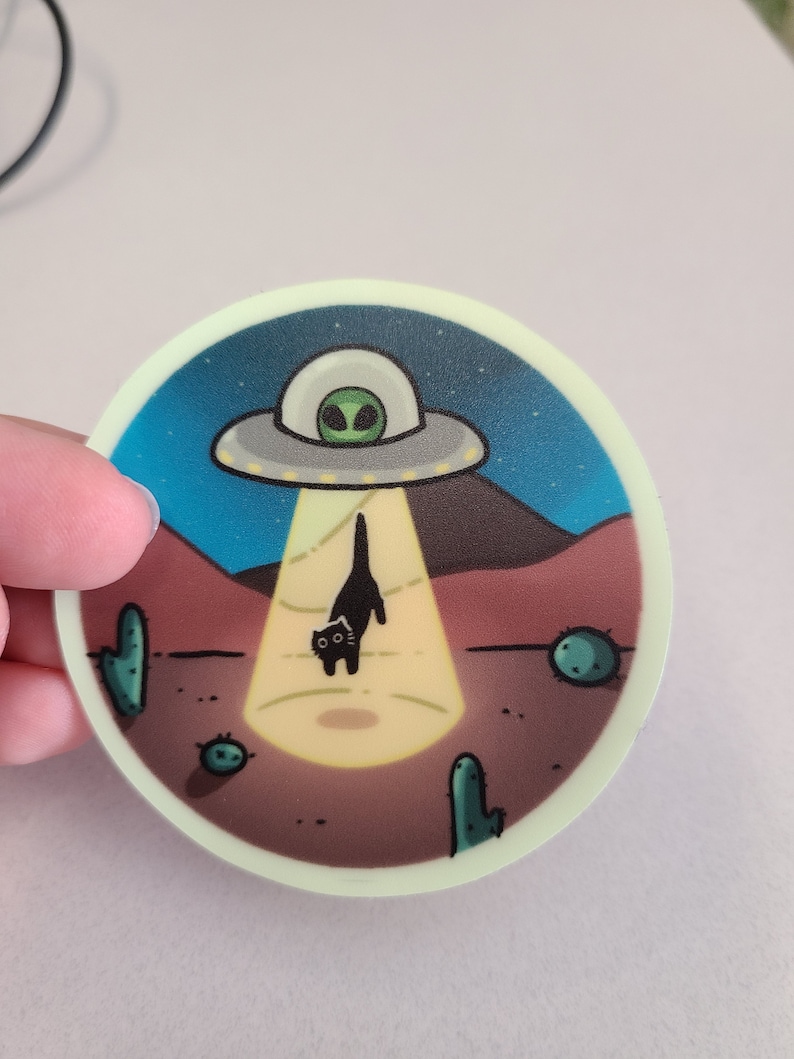 May include: A round sticker with a black cat standing in a beam of light from a flying saucer. The saucer is white with a green alien inside. The background is a desert scene with cacti.