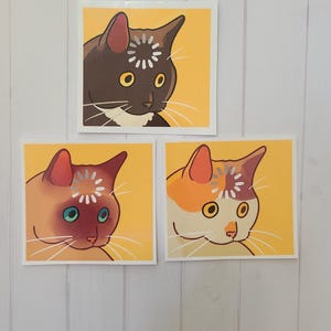 May include: Three square stickers featuring stylized cat portraits. Each cat has a different color scheme: brown, orange, and orange and white. Each cat has a floral design on its forehead and is set against a yellow background.