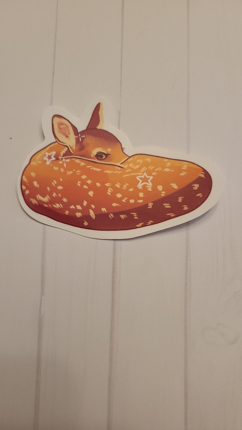 May include: A sticker featuring a curled-up fawn with a light brown coat and white spots. The fawn has a small head with large ears and a dark eye. Two white star shapes are on the fawn's body. The sticker has a white border.