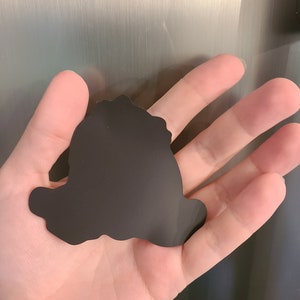Pu&ograve; includere: Silhouette nera di un personaggio dei cartoni animati, forse un cane, con una forma arrotondata e un design semplice. La silhouette &egrave; realizzata in un materiale nero flessibile ed &egrave; probabilmente una calamita.