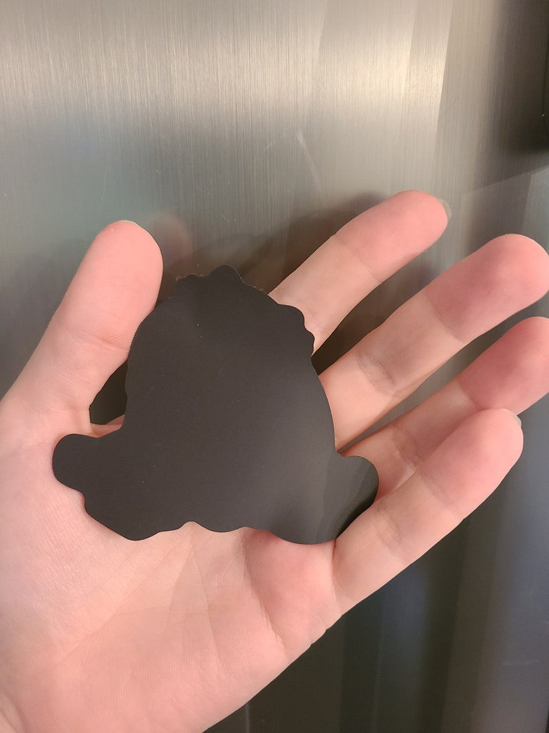 Pu&ograve; includere: Silhouette nera di un personaggio dei cartoni animati, forse un cane, con una forma arrotondata e un design semplice. La silhouette &egrave; realizzata in un materiale nero flessibile ed &egrave; probabilmente una calamita.