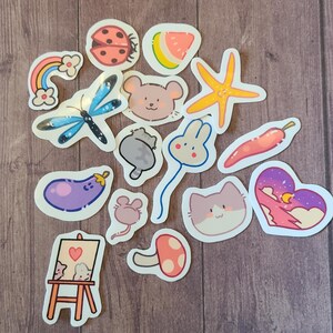 Op de afbeelding: Een verzameling kleurrijke stickers met verschillende schattige personages en objecten, waaronder een lieveheersbeestje, een watermeloen, een zeester, een muis, een konijn, een kat, een paddenstoel, een aubergine, een regenboog, een libel, een chilipeper en een hart met sterren en een maan.