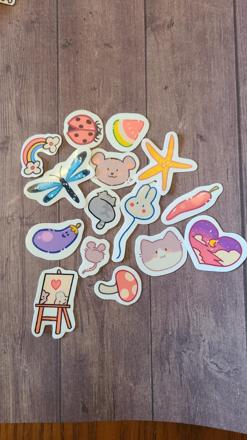 Op de afbeelding: Een verzameling kleurrijke stickers met verschillende schattige personages en objecten, waaronder een lieveheersbeestje, een watermeloen, een zeester, een muis, een konijn, een kat, een paddenstoel, een aubergine, een regenboog, een libel, een chilipeper en een hart met sterren en een maan.